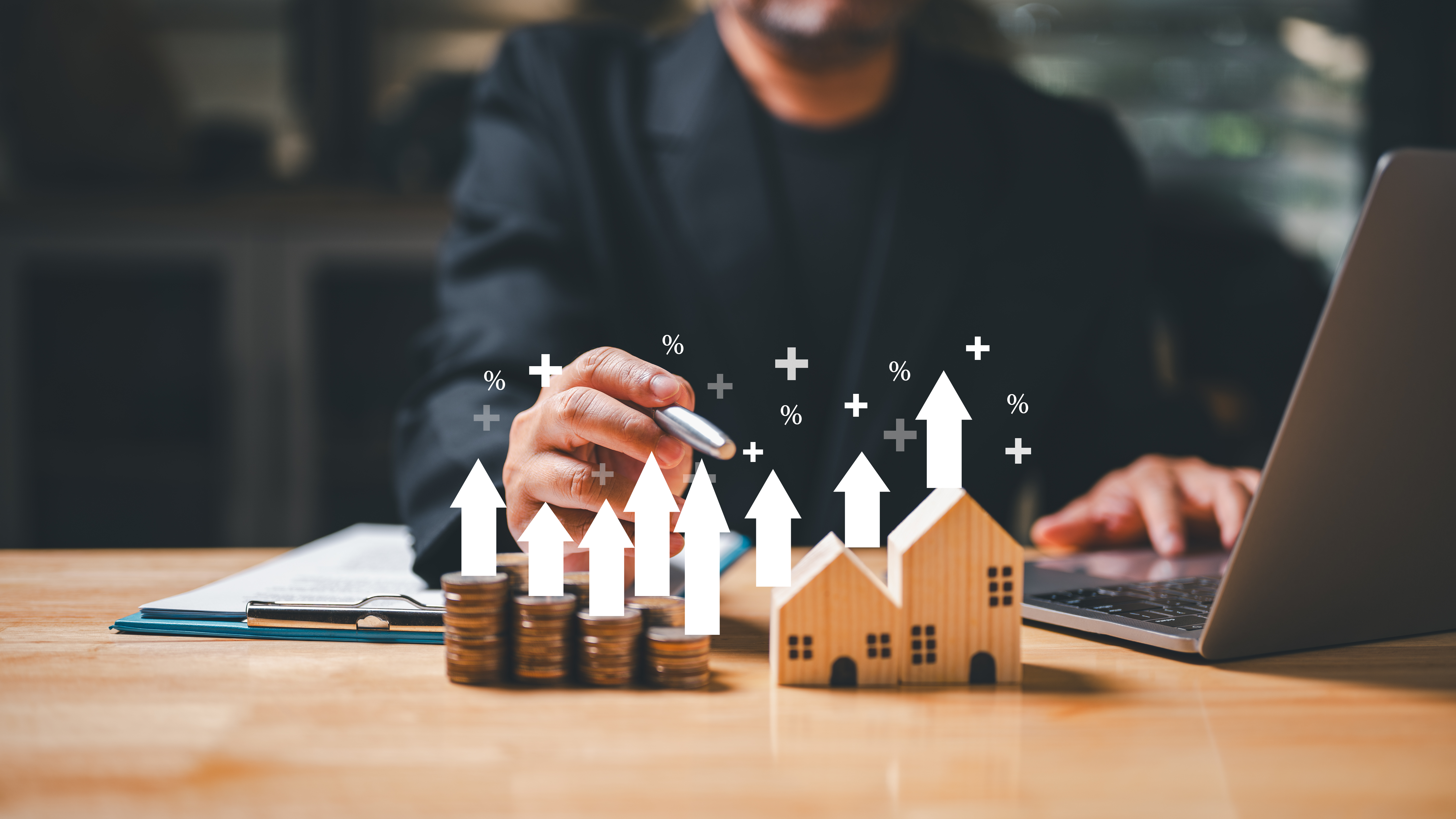 A person holding a pen pointing at little math symbols floating above a figurine of a house beside a stack of coins